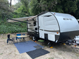 An EVO travel trailer, possibly an RV rental, set up at a leafy campsite with awning extended, chairs, and table.