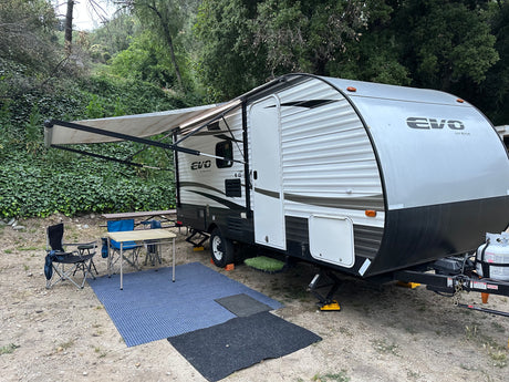 An EVO travel trailer, possibly an RV rental, set up at a leafy campsite with awning extended, chairs, and table.