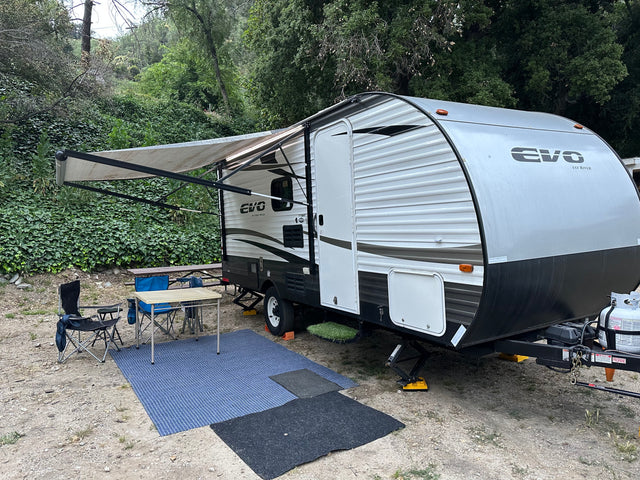 An EVO travel trailer, possibly an RV rental, set up at a leafy campsite with awning extended, chairs, and table.