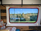Inside an RV rental, a window frames a tranquil river, trees, and lamppost. Cozy campervan interior with wood paneling and controls.