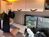 RV rental interior: overhead cabinets with leather pulls, a coat rack with hats, plush pillows, and an outdoor view.