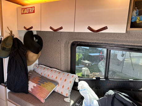 RV rental interior: overhead cabinets with leather pulls, a coat rack with hats, plush pillows, and an outdoor view.