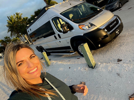 Smiling woman on a sandy beach next to her RV rental van, ready for adventure under the palms.