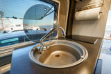 Inside an RV rental: compact kitchen with sink, faucet, stovetop, paper towels, and screened window.