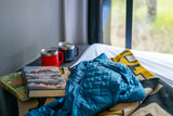 Enjoy your RV rental: two books, Yeti mugs, a warm jacket, and a cozy blanket beside a window with nature views.