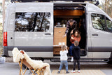 Family enjoys an RV rental: Dad gives treats to Mom in the campervan, while kids watch. Perfect for an outdoor family camping adventure.