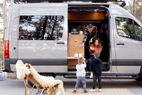 Family enjoys an RV rental: Dad gives treats to Mom in the campervan, while kids watch. Perfect for an outdoor family camping adventure.