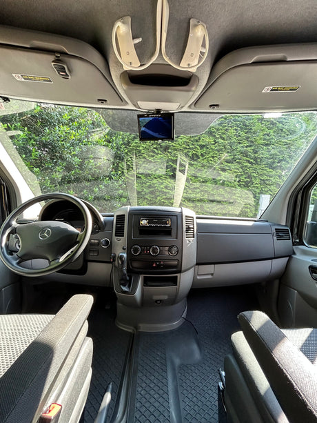 RV rental's front interior: Mercedes dashboard, steering wheel, and central console, with a clear view of green foliage outside.