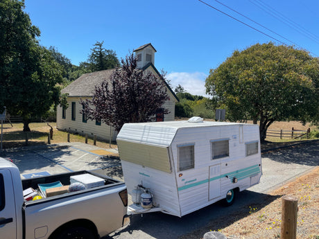 Vintage white and teal RV rental hitched to a truck, parked outside a historic church/schoolhouse on a sunny day.