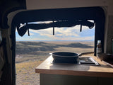 Inside an RV rental, a pan sits on a stovetop with olive oil nearby. The large window reveals a vast, hilly desert landscape.