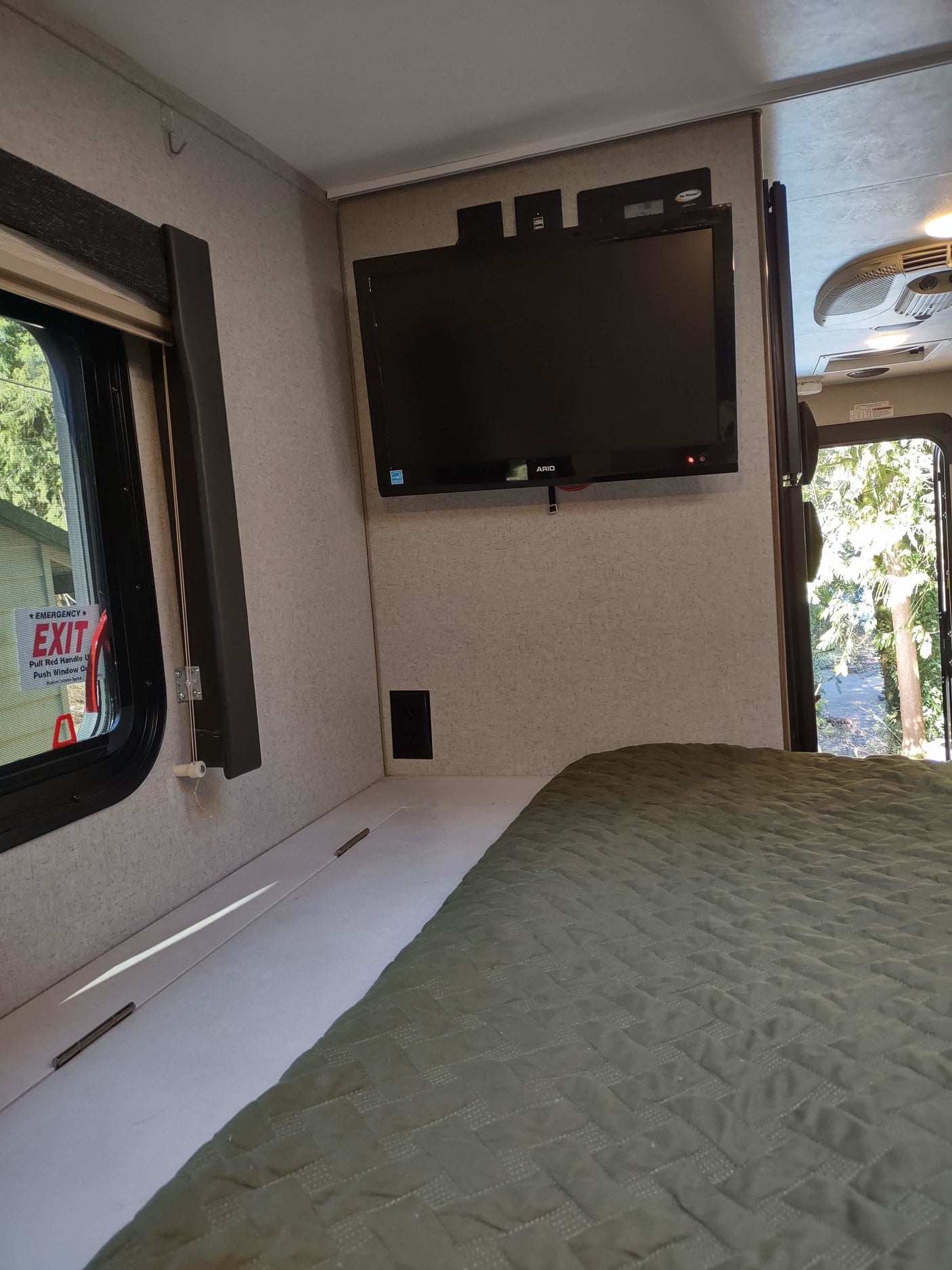 Cozy RV rental bedroom with a mounted TV, comfortable bed, emergency exit window, and a view to the outdoors.