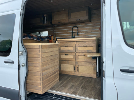 A white RV rental van interior with custom wooden kitchen, storage, bed, and paneling, designed for comfortable travel.