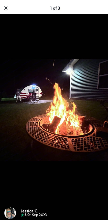 Night scene: Warm fire pit ablaze, with a white teardrop RV rental, chairs, and a person in the background. Cozy camping.