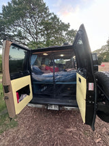 An RV rental camper van with open back doors. A person sleeps on a bed inside, a Hawaii license plate visible.