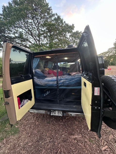 An RV rental camper van with open back doors. A person sleeps on a bed inside, a Hawaii license plate visible.
