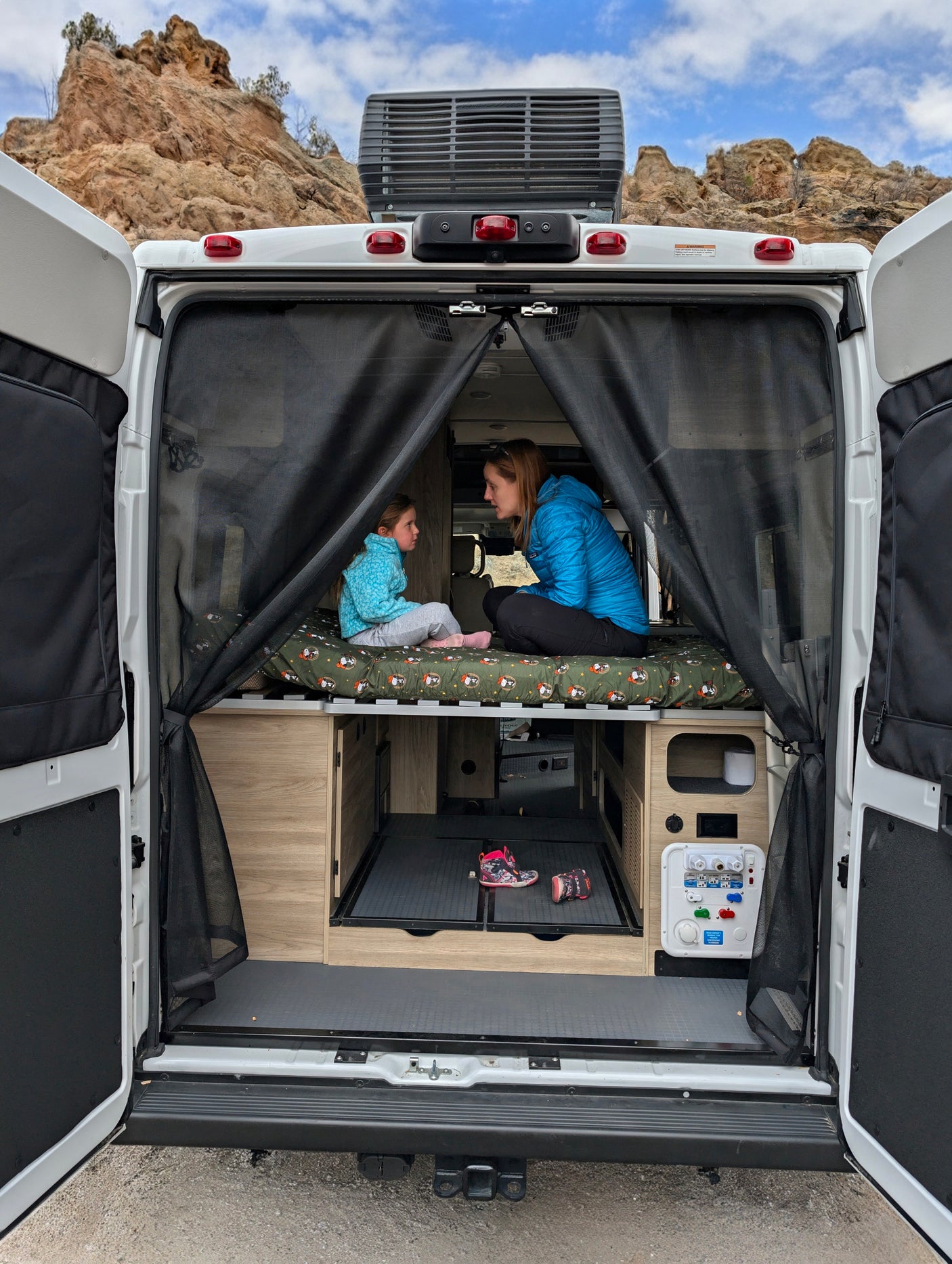 Rear of an rv rental with doors open, revealing a woman and child on a bed, facing scenic rocky mountains through mesh screens.