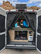 Rear of an rv rental with doors open, revealing a woman and child on a bed, facing scenic rocky mountains through mesh screens.