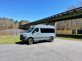 Light gray Mercedes Sprinter camper van (RV rental) by a muddy river & green bridge. Blue sky, winter trees, asphalt lot.