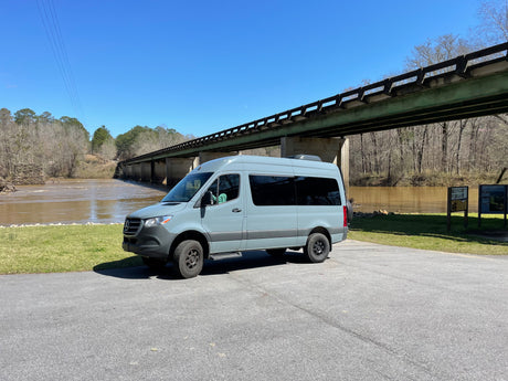 Light gray Mercedes Sprinter camper van (RV rental) by a muddy river & green bridge. Blue sky, winter trees, asphalt lot.