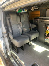 Inside an RV rental: two grey passenger seats with seatbelts, a kitchenette, and overhead storage nets.