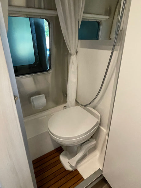 Compact RV rental bathroom featuring a white toilet, shower hose, curtain, and a slatted wooden floor mat.