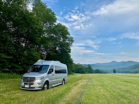 Silver Mercedes-Benz Sprinter RV rental parked in a green field, with mountains and trees under a blue sky, perfect for nature travel.