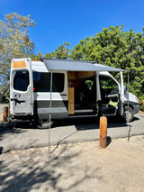 A white & gray converted camper van, excellent for RV rental, with an extended awning & open doors revealing its interior.