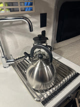 RV rental kitchen counter with a whistling kettle, French press, faucet, and roll-up dish rack, perfect for morning coffee.