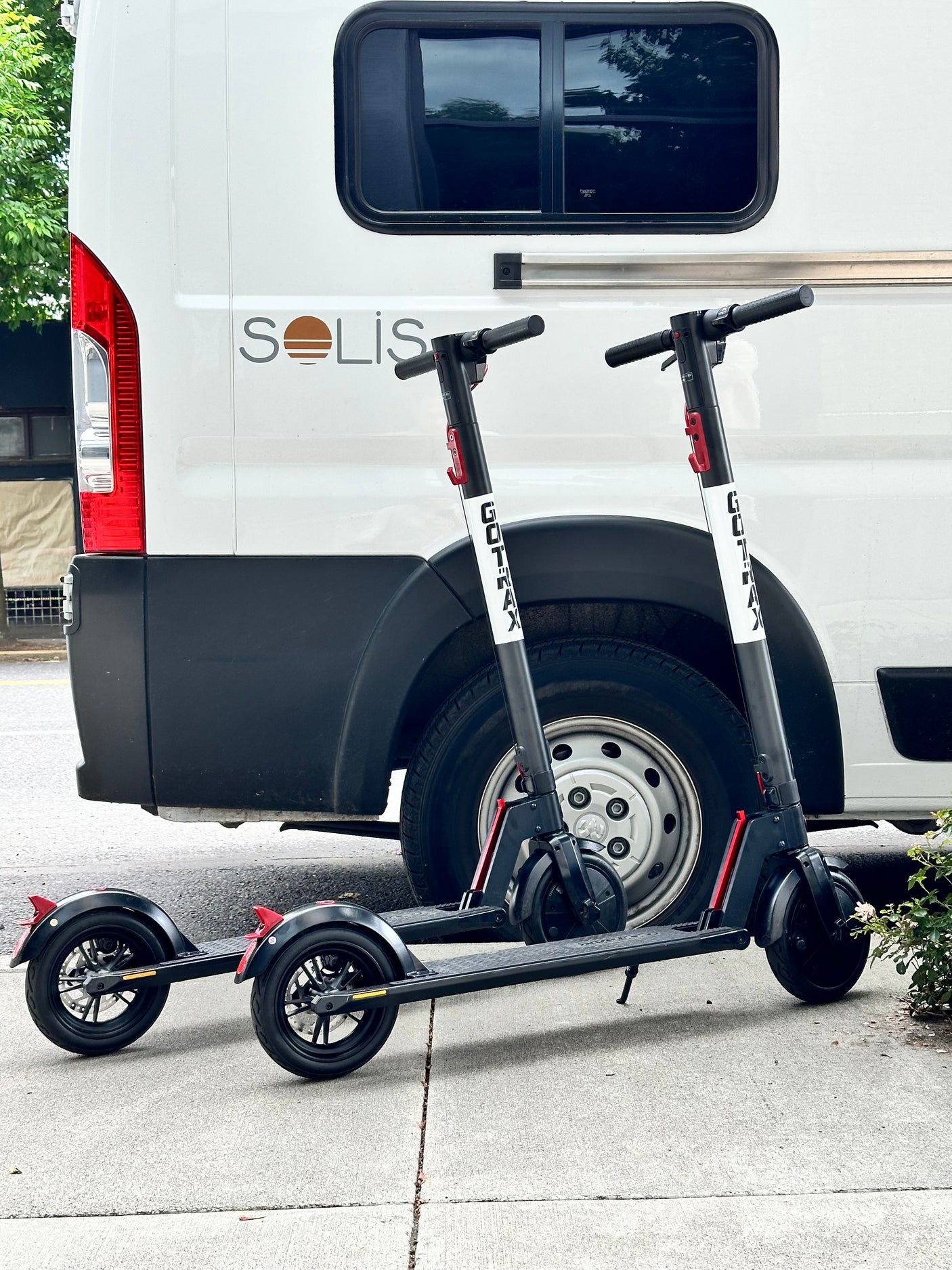 A white Solis RV rental van with two black GOTRAX electric scooters parked on the sidewalk.