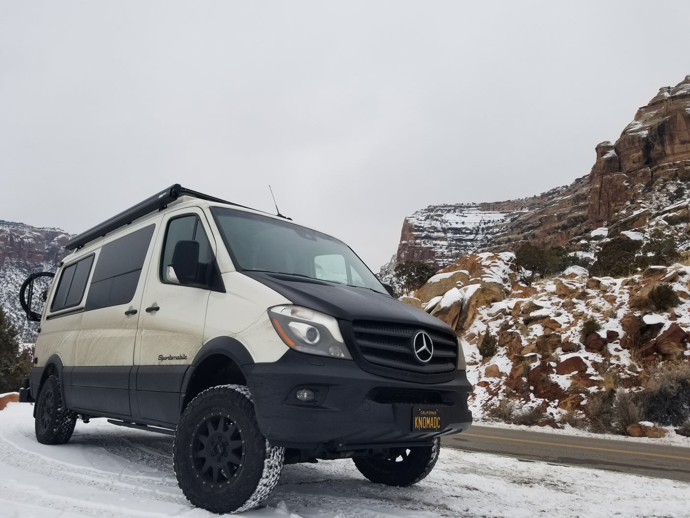 White and black Mercedes-Benz Sprinter Sportsmobile, an RV rental ready for snowy off-road adventures amidst rock formations.