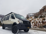 White and black Mercedes-Benz Sprinter Sportsmobile, an RV rental ready for snowy off-road adventures amidst rock formations.