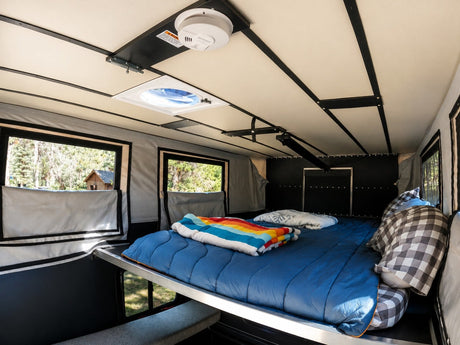 Bright rv rental interior showing a comfortable bed with a colorful blanket, windows with forest views, and a ceiling vent.