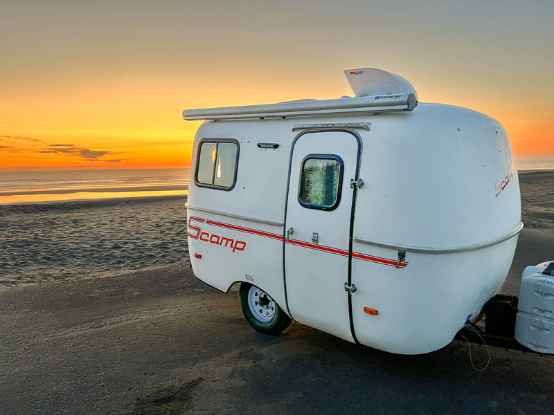 A white Scamp RV rental on a sandy beach at sunset. Vibrant orange and yellow sky over the ocean. Ideal for coastal camping. — photo 3
