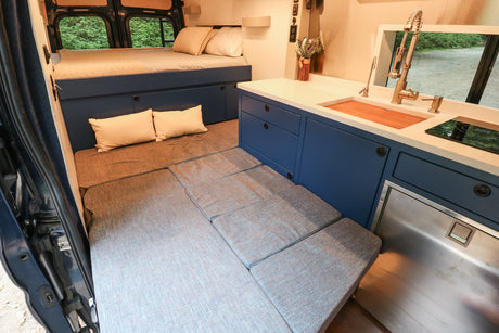 Modern RV rental interior showing a comfortable bed, convertible seating area, and a fully equipped kitchen with a sink & fridge.