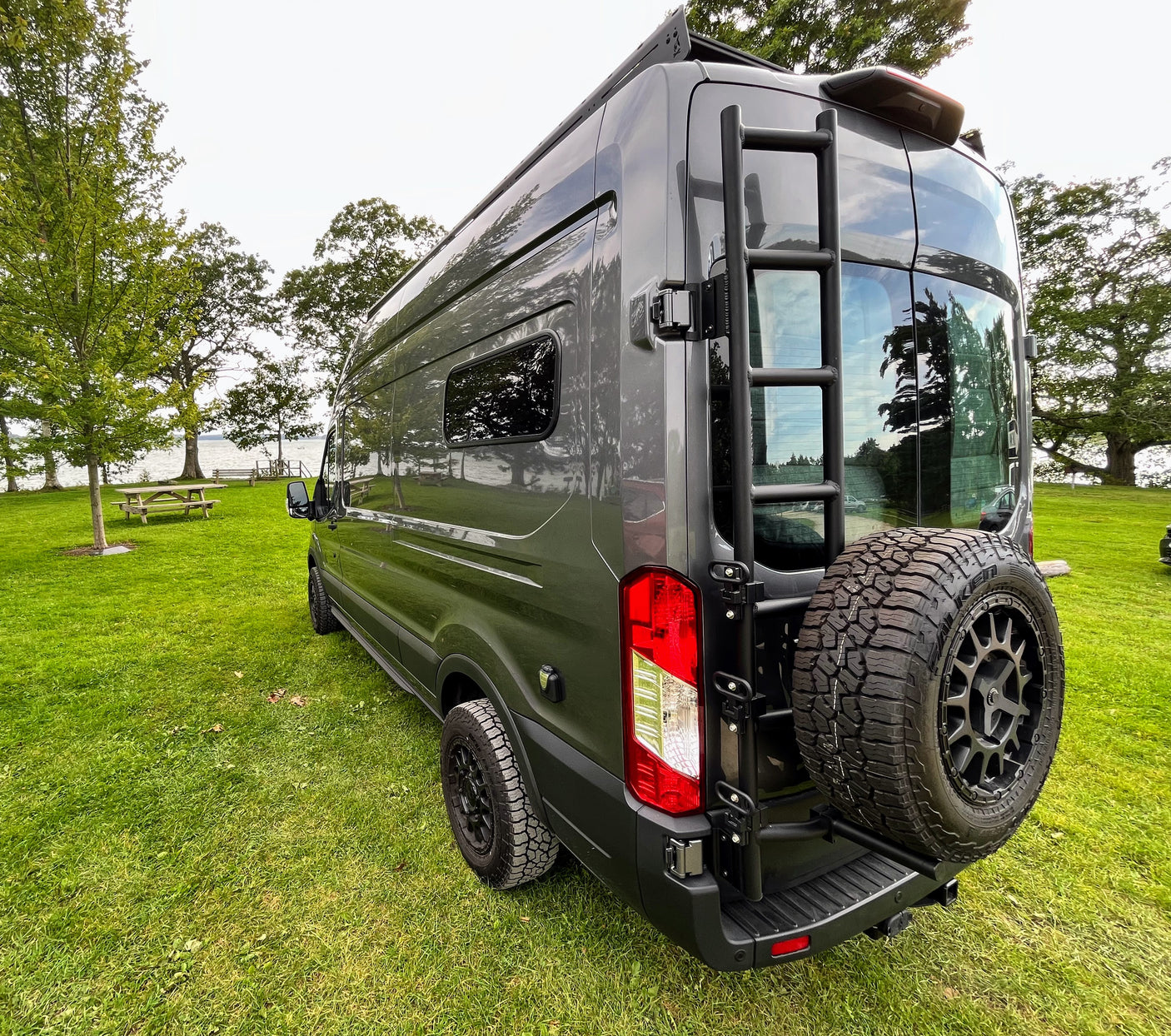 Stylish grey camper van parked on green grass by a lake, featuring a rear ladder & spare tire. Ideal for an rv rental adventure.