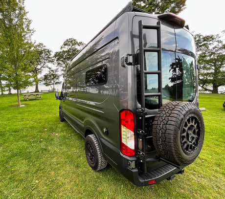 Stylish grey camper van parked on green grass by a lake, featuring a rear ladder & spare tire. Ideal for an rv rental adventure.