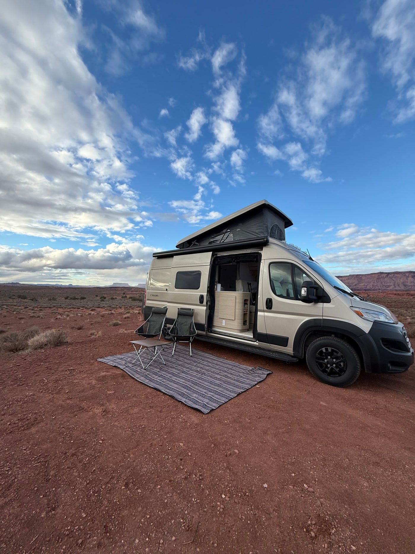 Beige RV rental parked on red desert earth with a pop-top extended, chairs, and a mat out for camping. — photo 6