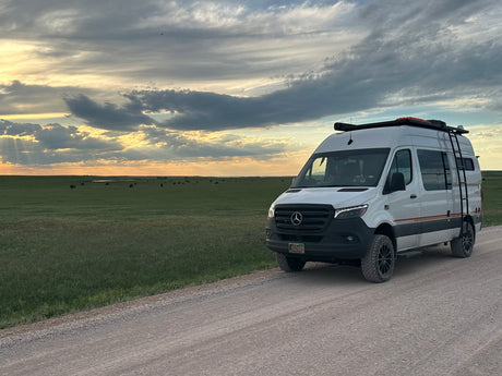 White Mercedes Sprinter RV rental on a dirt road at sunset. Explore vast green plains with distant bison from your van.