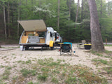 Teardrop RV rental ready for adventure at a gravel forest campsite, featuring an open kitchenette.
