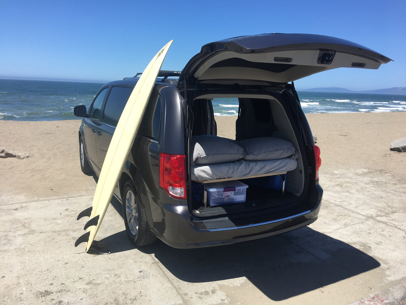 Dark minivan converted into a camper with a bed, surfboard, and ocean view. Perfect for an RV rental adventure.