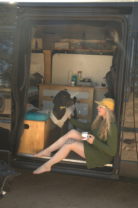 Woman & dog relax in a custom RV rental. Barefoot in the doorway, she sips coffee; the van reveals a cozy kitchen.