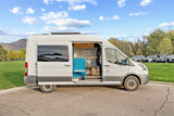 White rv rental camper van with open side door reveals a custom blue and wood interior. Parked near mountains on a sunny day.