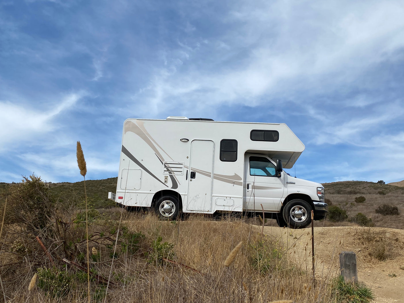 White RV rental parked on a dirt path in dry, grassy hills under a blue sky, perfect for outdoor adventures.
