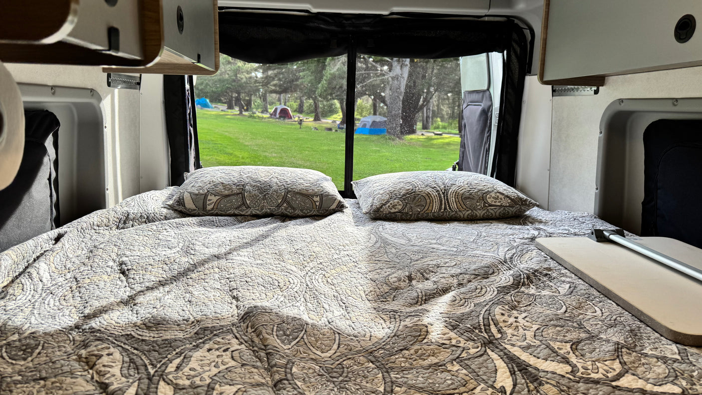 Inside an RV rental, a comfy bed faces a large window overlooking a green campsite with tents and trees outside.