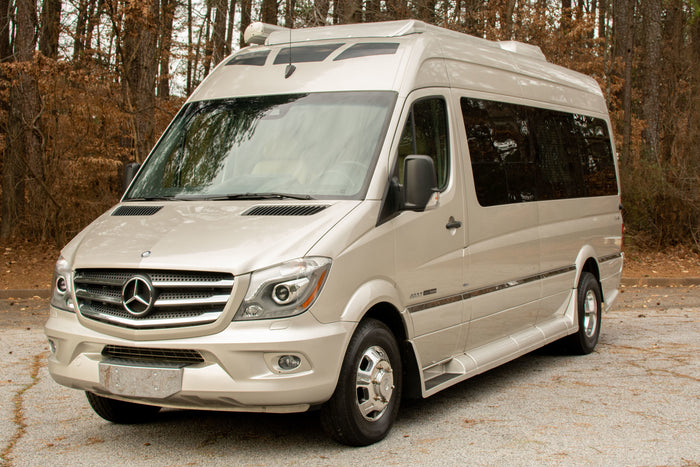 This champagne Mercedes-Benz Sprinter RV, ideal for an RV rental, is parked outdoors amidst trees.