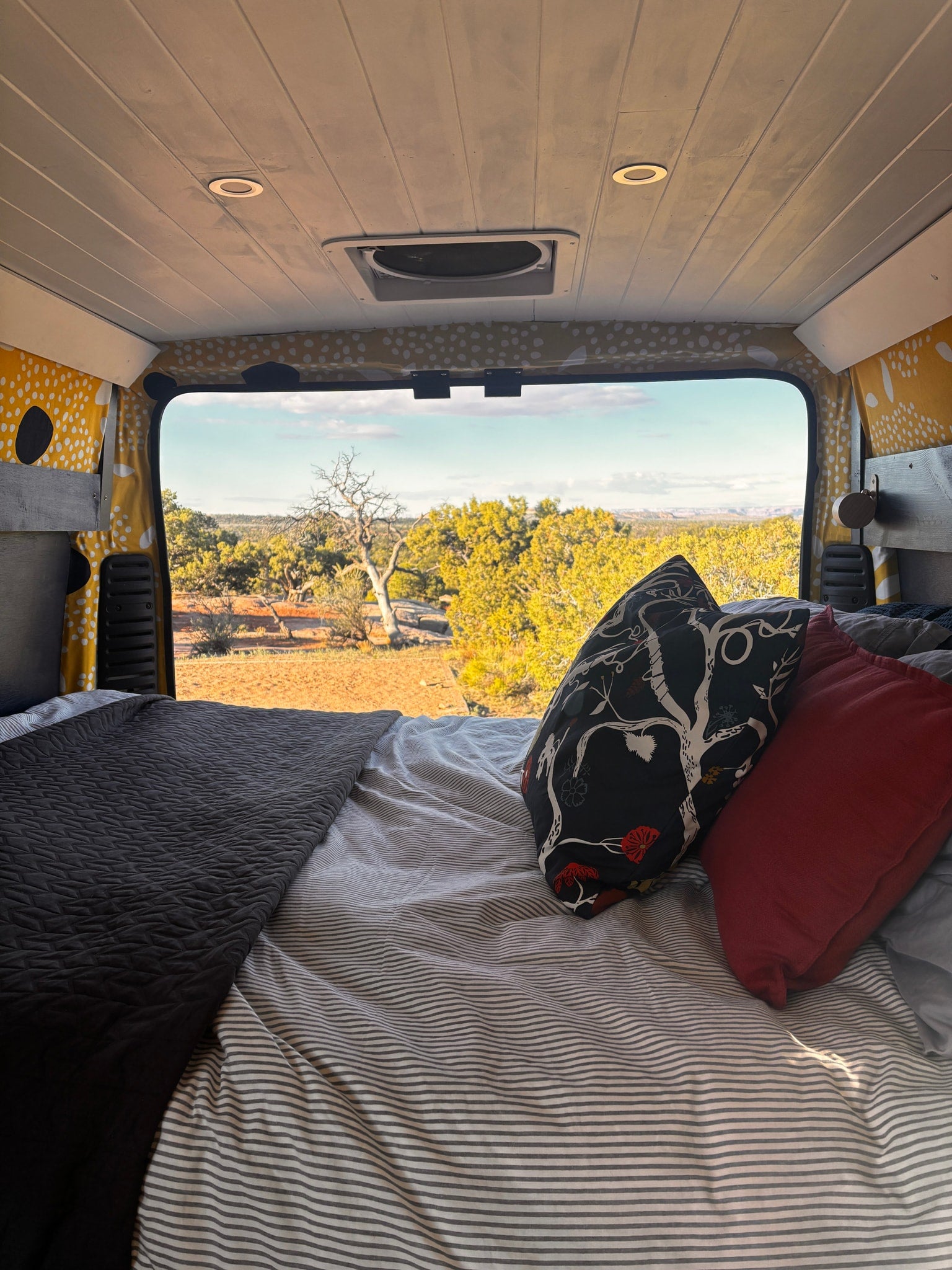 Cozy rv rental bed with striped sheets, decorative pillows, and a bright desert landscape view from the back window.