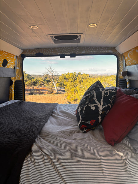Cozy rv rental bed with striped sheets, decorative pillows, and a bright desert landscape view from the back window.