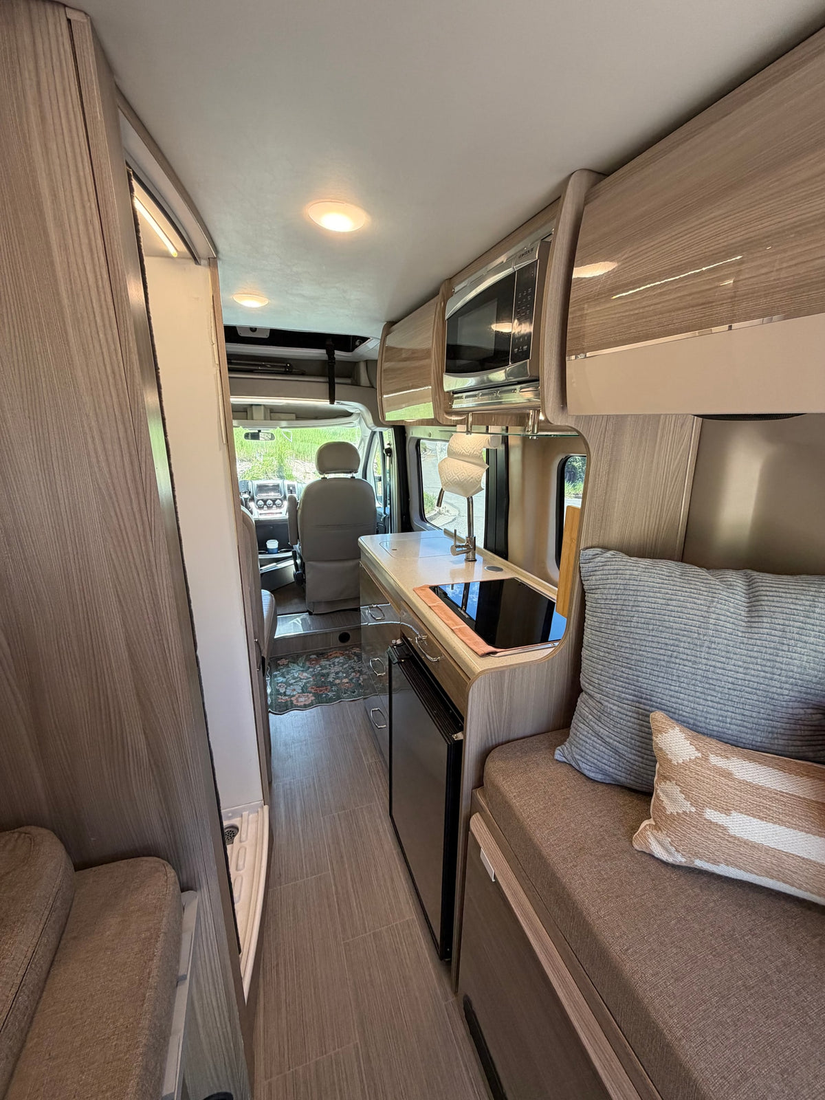 Modern RV rental interior featuring a compact kitchen with microwave, sink, and stovetop, plus comfy seating and the driver's cabin. — photo 7