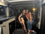 Two smiling women inside their RV rental camper van. Features a compact kitchen and seating, perfect for mobile travel.