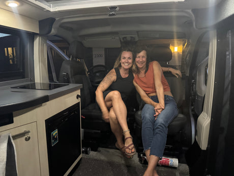 Two smiling women inside their RV rental camper van. Features a compact kitchen and seating, perfect for mobile travel.